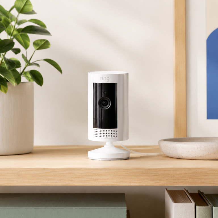 Ring Indoor Cam in white sitting on a light wood shelf in a bright minimalist room with a plant and neutral decor
