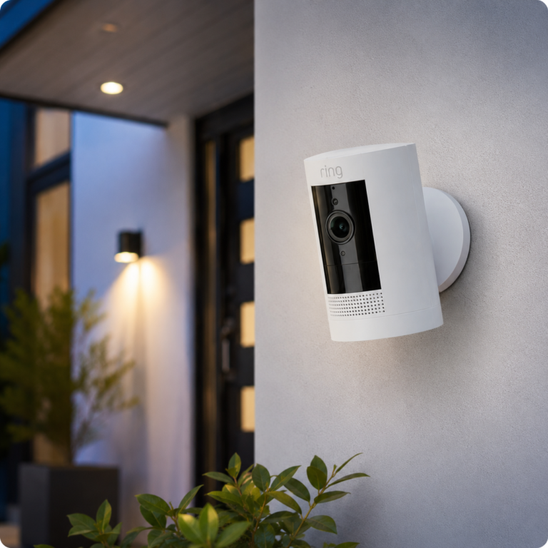 Ring Outdoor Cam mounted on a light exterior wall near a modern front door with greenery in the foreground